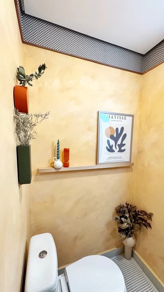 Lavabo pequeno com pintura amarela na parede, vaso de planta na parede e no chão, detalhe xadrez.