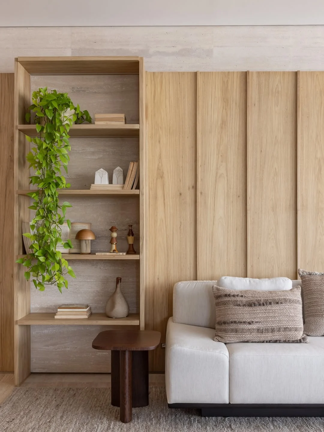 Sofá claro com almofada marrom texturizada, painel de madeira e planta no fundo.