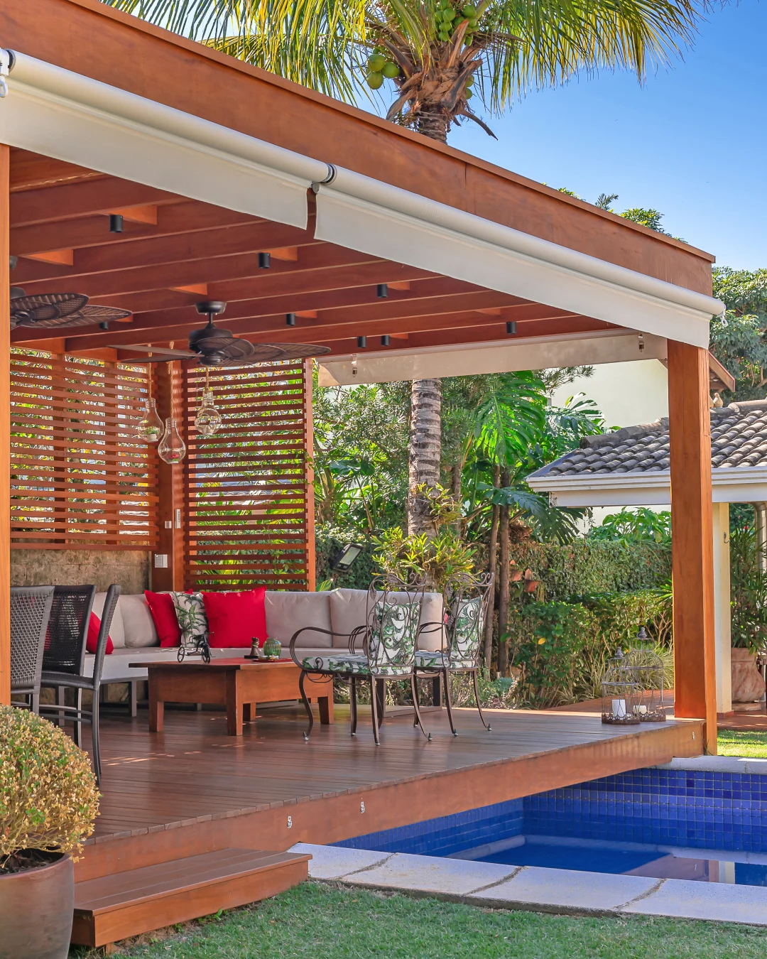Área externa com pergolado de madeira, sofá neutro e almofadas vermelhas em destaque.