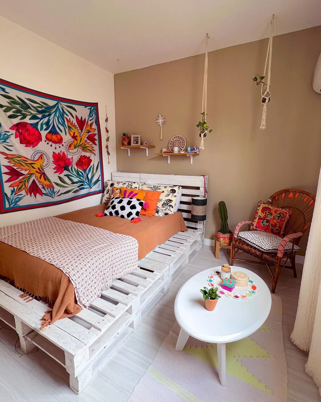 Cama com pallet branco, pano estampado na parede, mesa de centro branca e almofada preto e branco na cama.