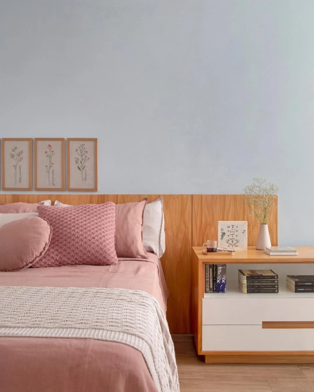 Quarto adulto com cabeceira de madeira, quadros de flores e almofadas rosadas.