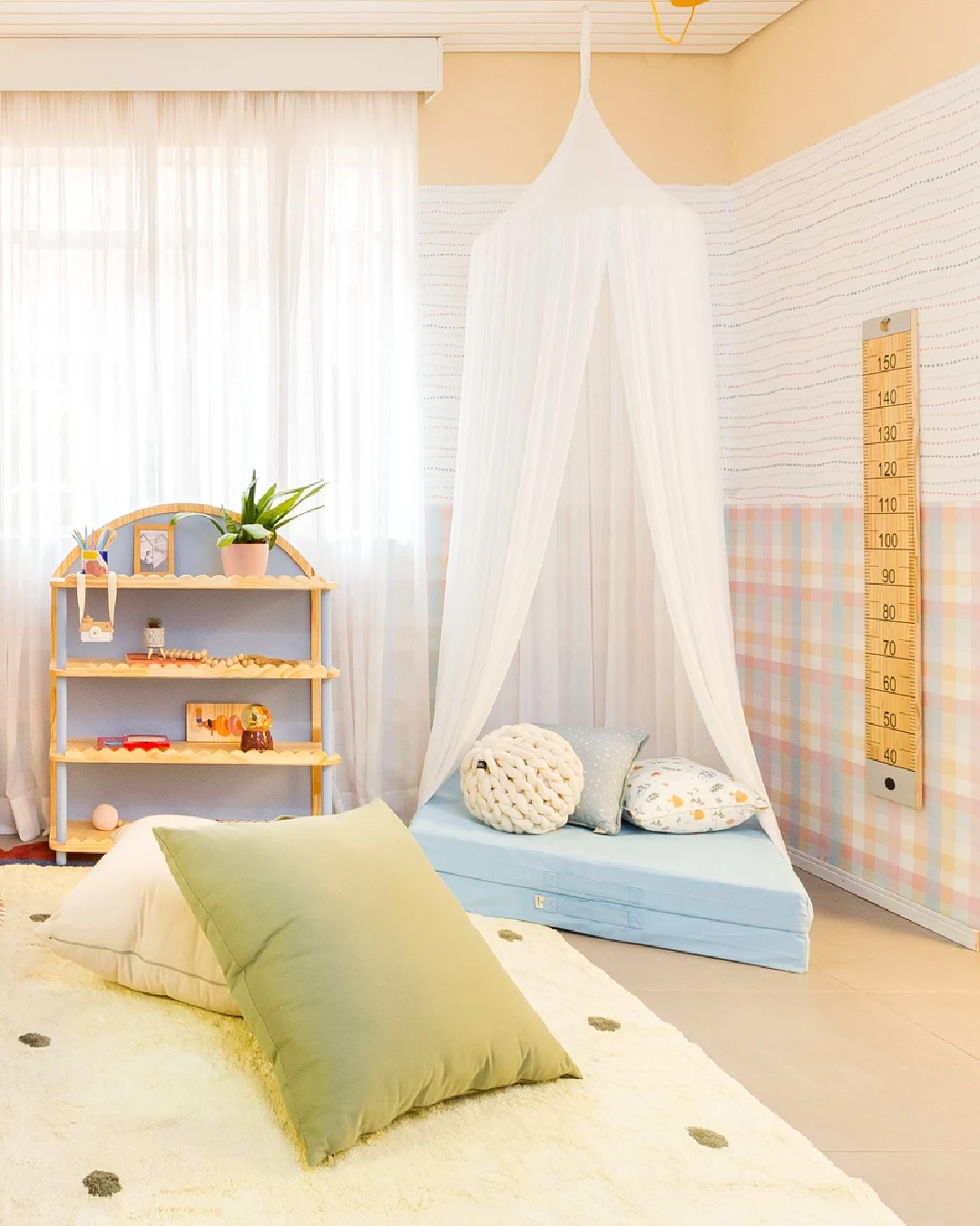 Quarto infantil com cantinho para brincar cheio de almofadas, fita métrica gigante na parede e estante baixa.