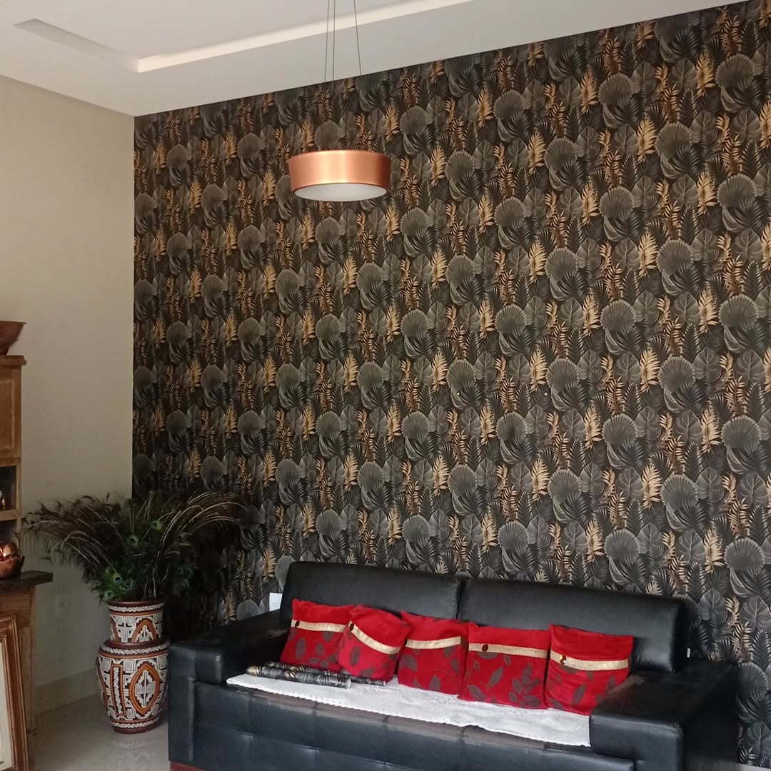 Parede com papel adesivo chamativo em tons de preto e dourado, sofá preto com almofadas vermelhas e planta.