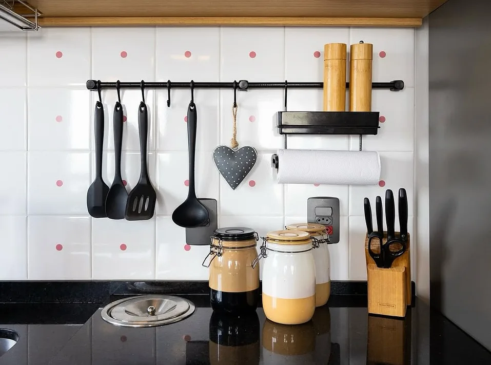 Cozinha com azulejo branco e pontinho rosa, utensílios pretos pendurados.